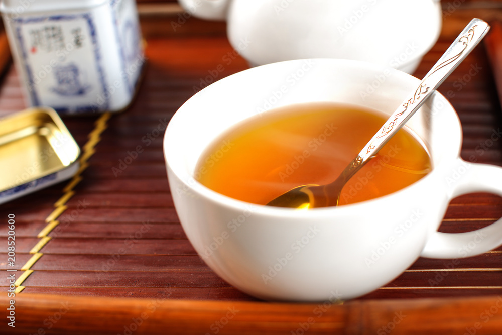 White porcelain cup of hot steaming tea, with spoon, placed on bamboo tray. White porcelain cup of hot steaming tea, with spoon, placed on bamboo tray.