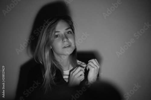 Attractive young  female actress wearing black fur jacket looking tenderly at camera in closed posture, keepings arms on breast, standing against white wall during audition under light of soffits.