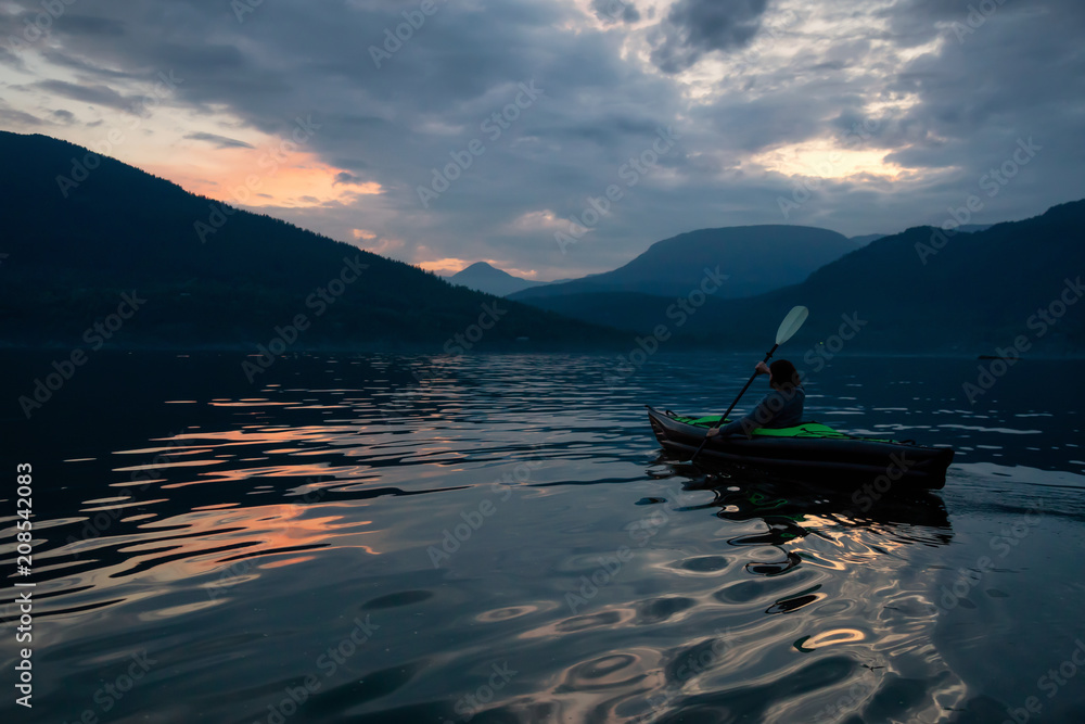 Adventurous woman kayaking during a vibrant sunset surrounded by Canadian Mountain Landscape. Taken in Howe Sound, North of Vancouver, BC, Canada.