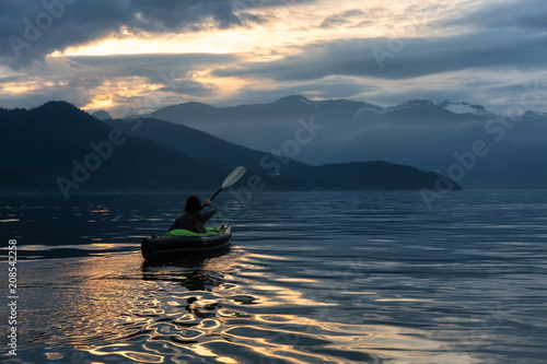 Adventurous woman kayaking during a vibrant sunset surrounded by Canadian Mountain Landscape. Taken in Howe Sound, North of Vancouver, BC, Canada.