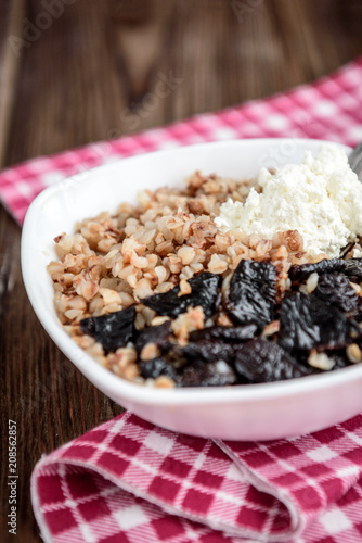 Boiled buckwheat with milk, prune and cottage cheese in white bowl on dark wooden table
