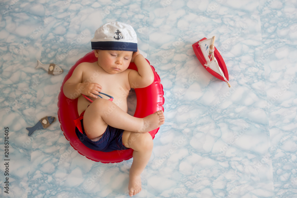 Cute baby boy sleeping on a tiny inflatable swim ring wearing swimsuit shorts and sunglasses. Stock Adobe Stock
