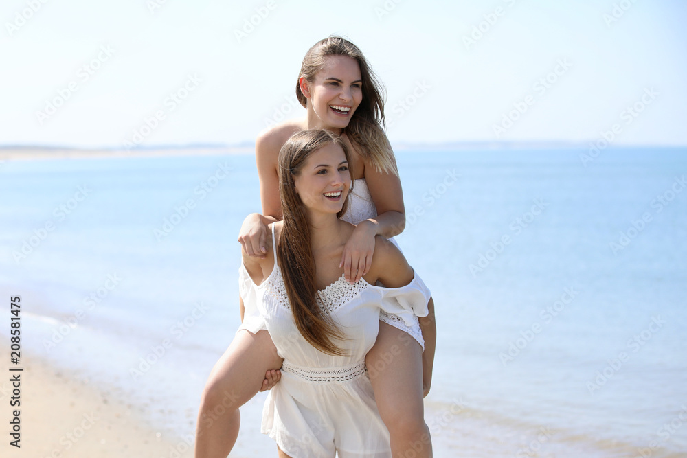 Zwei hübsche junge Frauen an einem Strand schauen in eine Richtung, eine Frau wird Huckepack getragen