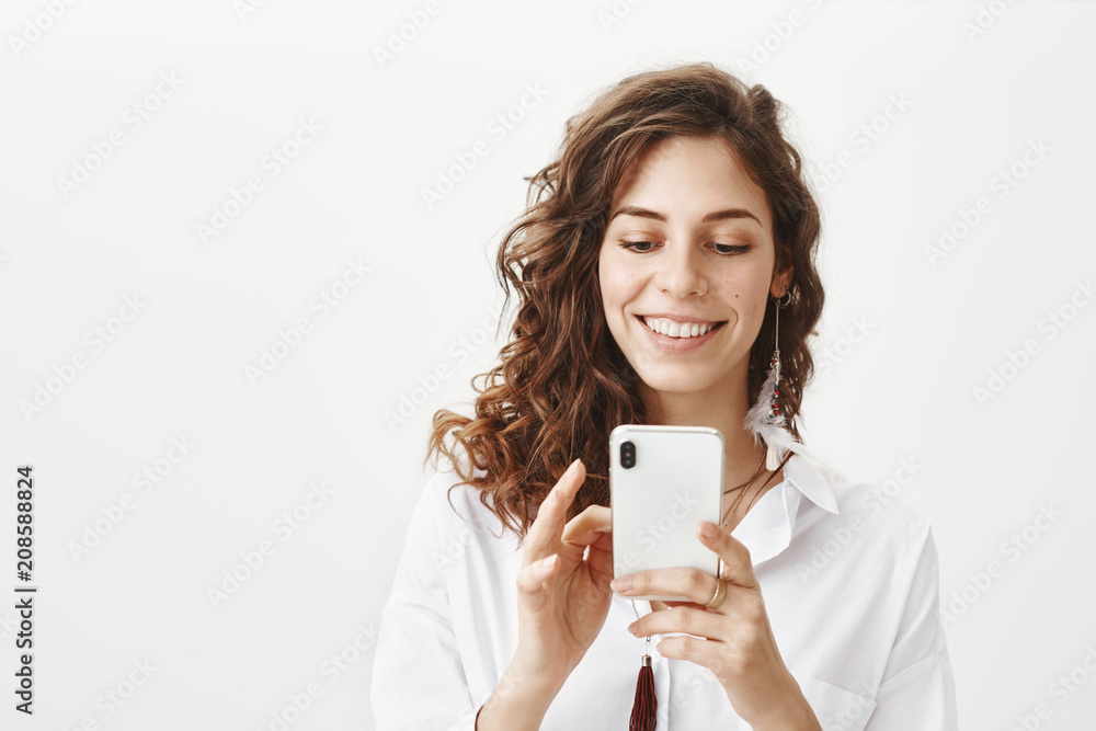 Indoor portrait of happy charming woman with feather earring typing something in smartphone while smiling broadly and being in good mood, standing over gray background. Girl plays game while on lunch Indoor portrait of happy charming woman with feather earring typing something in smartphone while smiling broadly and being in good mood, standing over gray background. Girl plays game while on lunch