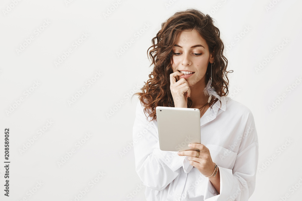 Portrait of curious intrigued beautiful woman biting finger while looking at tablet with excitement, standing over gray background. Girl thinks whether she want sent message or not Portrait of curious intrigued beautiful woman biting finger while looking at tablet with excitement, standing over gray background. Girl thinks whether she want sent message or not