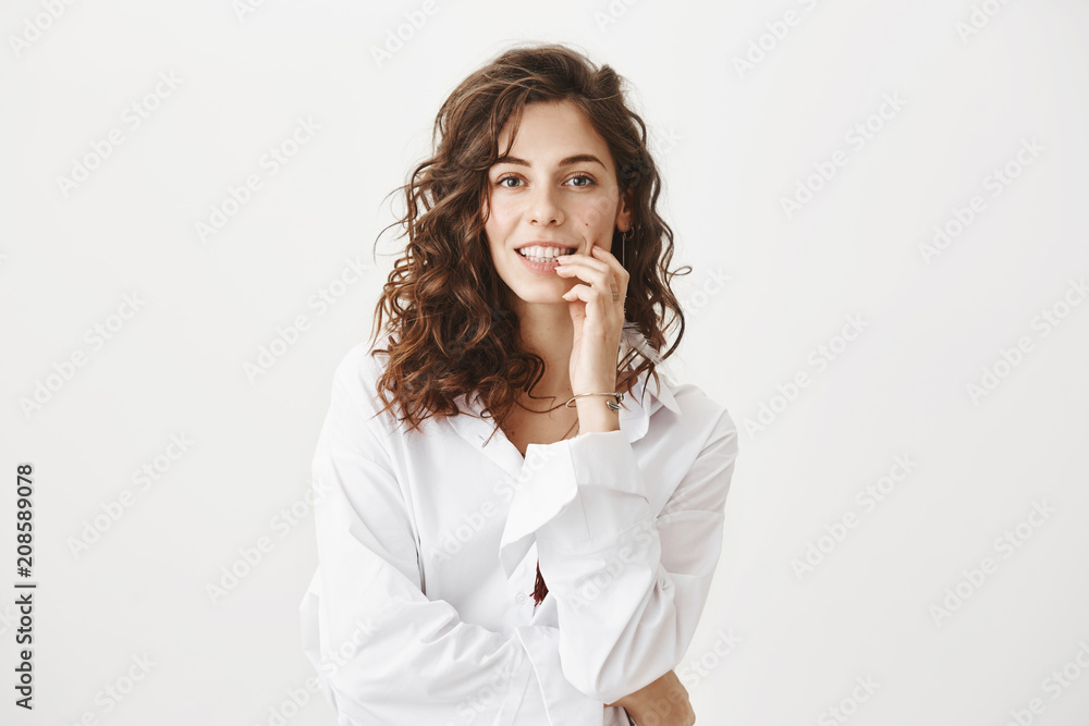 Charming adult caucasian woman holding fingers on lip while smiling and looking seductively at camera, being in romantic mood and flirting while standing against gray background. Charming adult caucasian woman holding fingers on lip while smiling and looking seductively at camera, being in romantic mood and flirting while standing against gray background.
