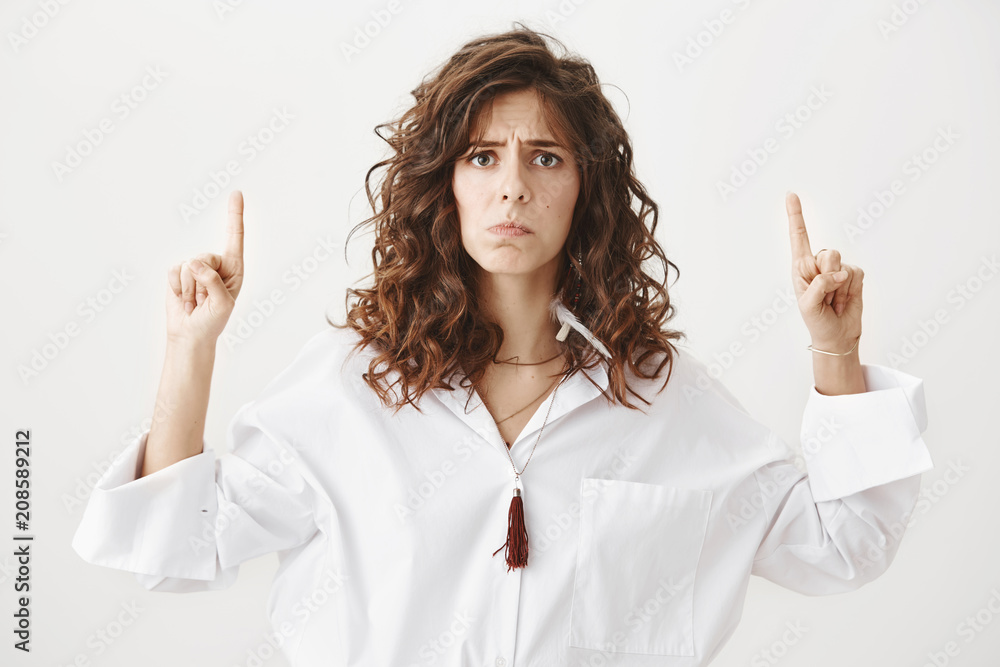 Gloomy upset charming caucasian female model pointing up and frowning, being offended or expressing bad mood, standing against gray background. Woman is sad and is about to cry or complain Gloomy upset charming caucasian female model pointing up and frowning, being offended or expressing bad mood, standing against gray background. Woman is sad and is about to cry or complain
