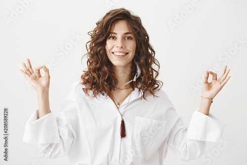 Positive joyful caucasian adult female with curly hair smiling and standing in meditating pose with zen gestures, being calm and confident over gray background. Girl tries to boost her mood with yoga