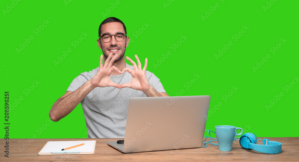 Happy young man doing a gesture of love sitting at his desk - Green background Happy young man doing a gesture of love sitting at his desk - Green background