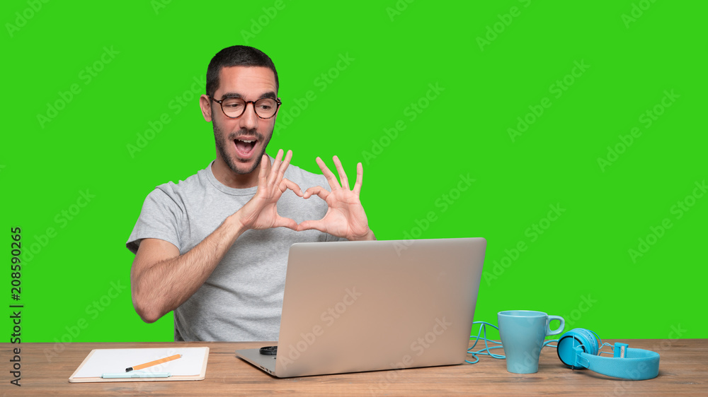 Happy young man doing a gesture of love sitting at his desk - Green background Happy young man doing a gesture of love sitting at his desk - Green background