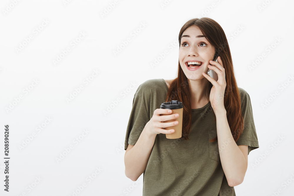 Woman sharing with friend during mobile talk with positive news. Studio shot of overwhelmed happy young girl, holding cup of coffee, talking on smartphone and looking up with dreamy cheerful look Woman sharing with friend during mobile talk with positive news. Studio shot of overwhelmed happy young girl, holding cup of coffee, talking on smartphone and looking up with dreamy cheerful look