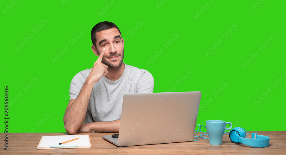 Concerned young man sitting at his desk and doing a gesture of observe - Green background