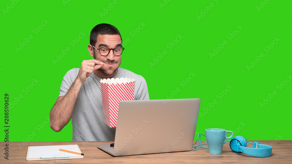 Surprised young man watching a film on his laptop and eating popcorn - Green background Surprised young man watching a film on his laptop and eating popcorn - Green background