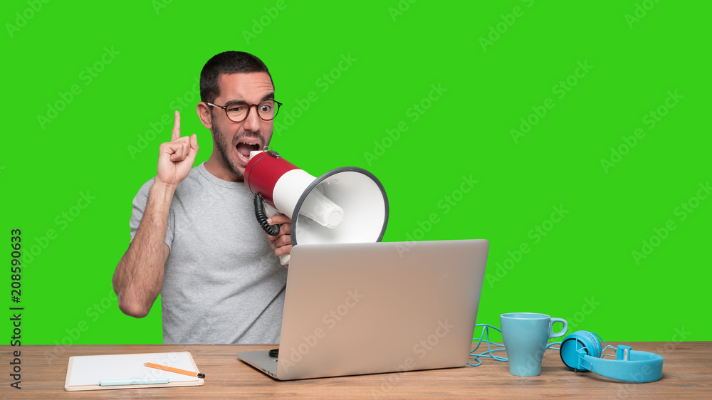 Young man sitting at his desk and using a megaphone - Green background Young man sitting at his desk and using a megaphone - Green background