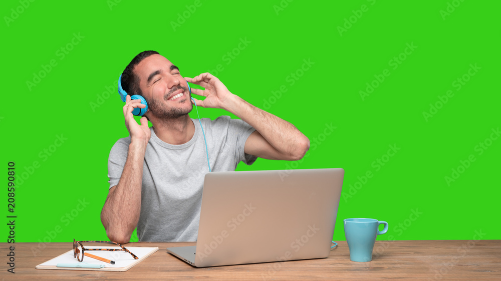 Happy young man sitting at his desk and using a headphones - Green background Happy young man sitting at his desk and using a headphones - Green background