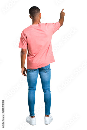 Standing young african american man pointing back with the index finger presenting a product from behind on isolated white background