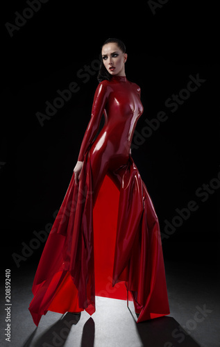 Brunette woman in red latex slim suit posing on dark background © FlexDreams