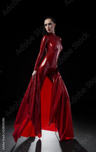 Brunette woman in red latex slim suit posing on dark background