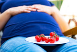 Pregnant woman with baby belly sitting in the sunbed with a plate with fresh tomatos on it on the legs