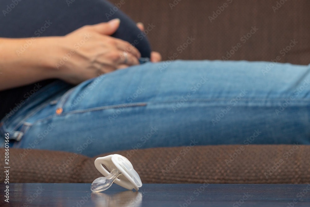 In front of a pregnant woman who is sitting on the sofa with her baby belly a soother is lying on a brown wooden table