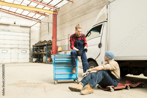 Young bearded male car technician chatting with his colleague while resting near broken truck in large service garage