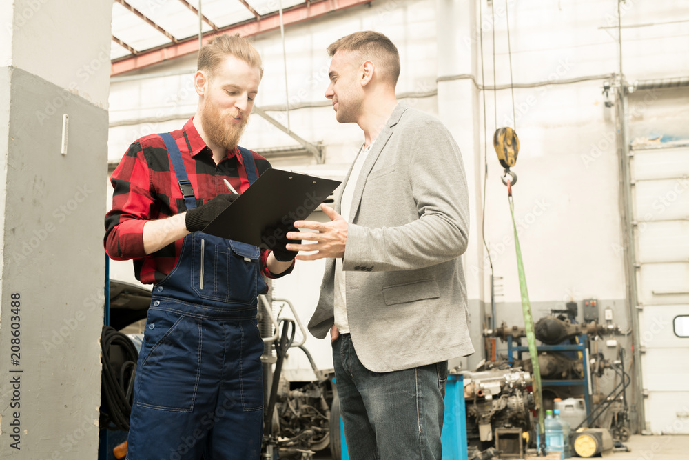 Young bearded auto mechanic looking at checklist and smiling while male customer thanking him for maintenance in auto repair service