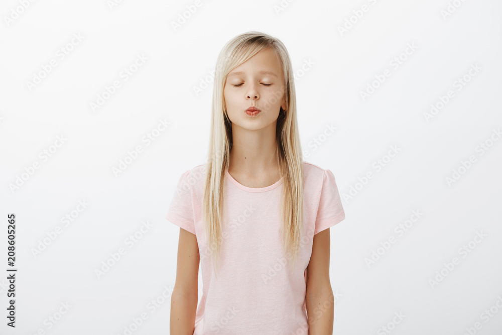 Girl learning how to kiss on tomatoes. Portrait of relaxed and focused fashionable child with blond hair, closing eyes and folding lips, giving mwah while standing against gray background Girl learning how to kiss on tomatoes. Portrait of relaxed and focused fashionable child with blond hair, closing eyes and folding lips, giving mwah while standing against gray background