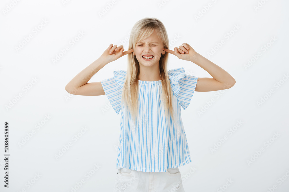 Fototapeta premium Girl hate when parents arguing. Displeased annoyed young child with blond hair in blue blouse, covering ears with index fingers and grimacing, hearing annoying sound, standing over gray wall