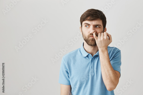 Can I dig to brain. Studio shot of clueless untidy caucasian guy looking aside with confused expression, picking in nose with pinky, thinking nobody sees him over gray background.