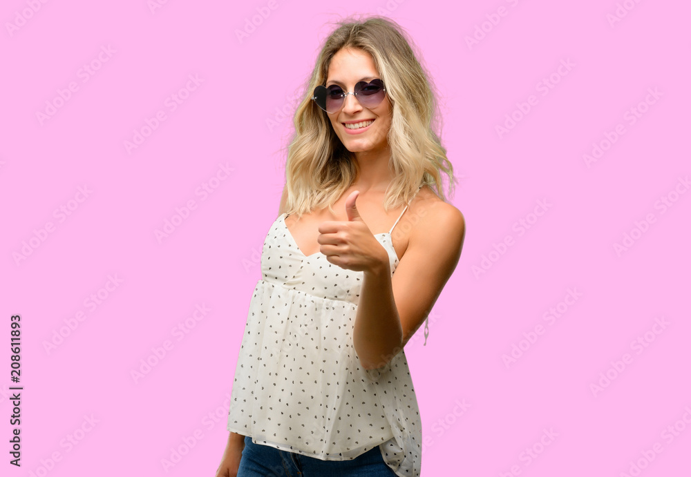 Young woman wearing sunglasses with heart shape smiling broadly showing thumbs up gesture to camera, expression of like and approval