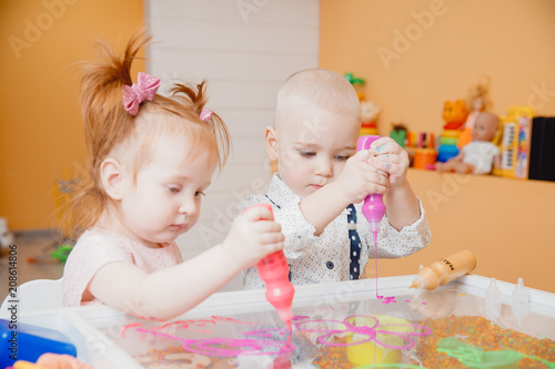 Close-up of baby man and girl hand holds liquid paint in bottle of purple, drawings on table.