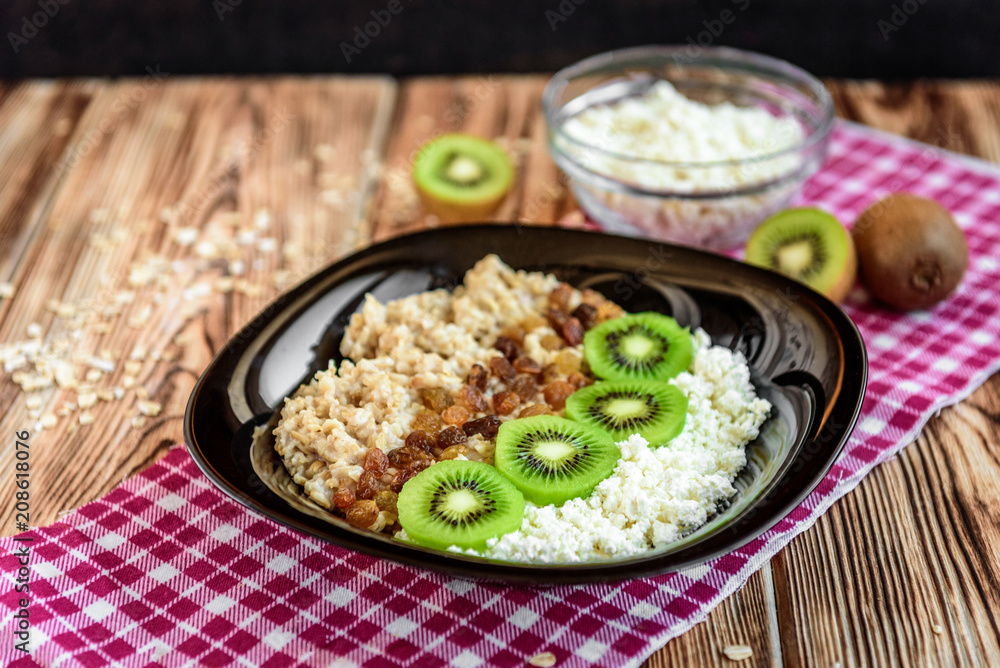Breakfast with oatmeal porridge, kiwi, cottage cheese and raisin on black plate on dark wooden background. Healthy breakfast and eating concept. Top view. 