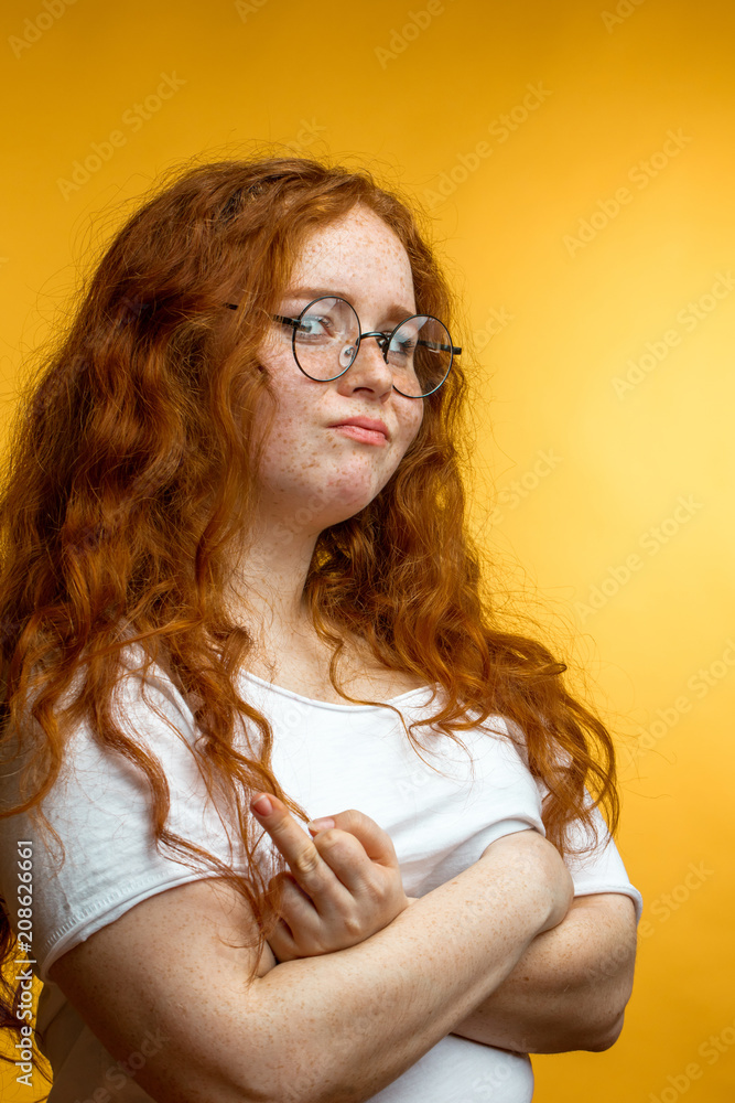 Negative human facial expression. Ginger long haired girl with arms crossed  over breast pouting lips and looking at camera with irritated and offended  face expression while standing isolated yellow Photos