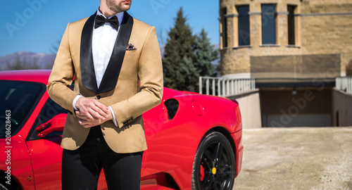 Man in custom tailored tuxedo, suit posing outdoors in front of expensive car photo