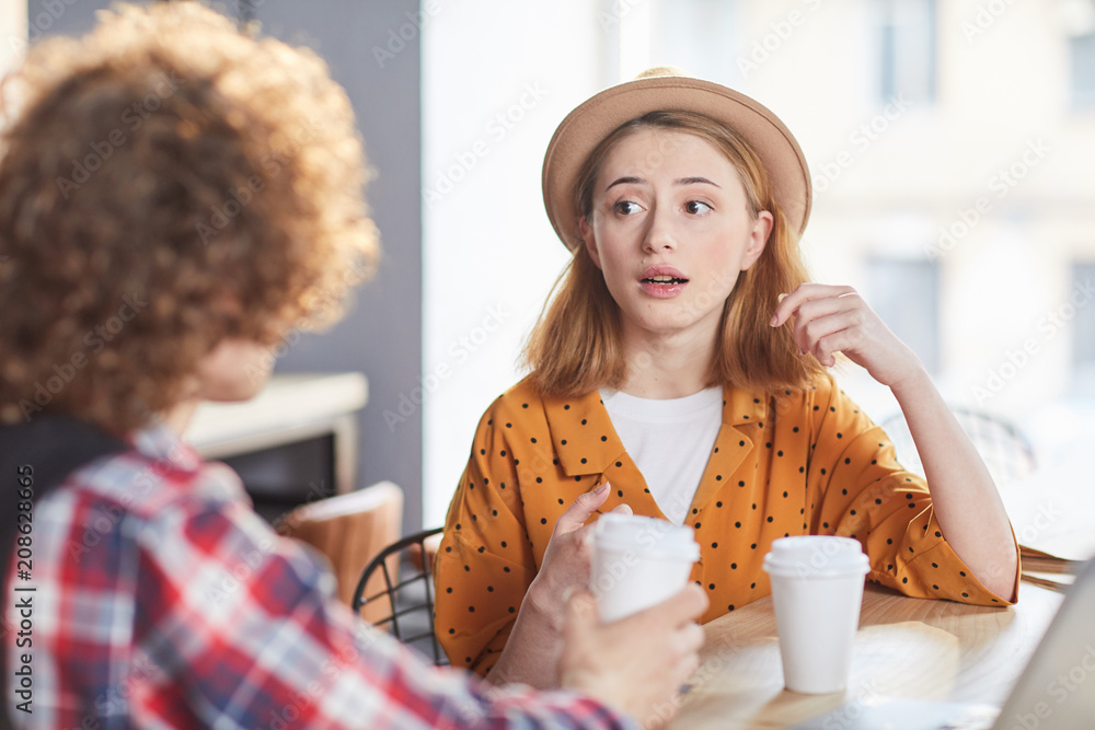 Young businesswoman in hat and yellow blouse explainign her colleague some working points during break in cafe Young businesswoman in hat and yellow blouse explainign her colleague some working points during break in cafe