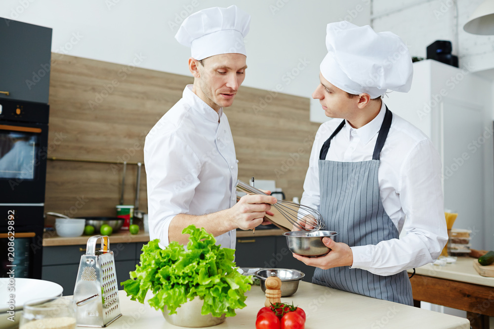 Chef with whisk mixing raw eggs in steel bowl while consulting his trainee in the kitchen Chef with whisk mixing raw eggs in steel bowl while consulting his trainee in the kitchen
