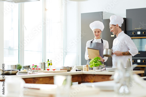 Young female making notes in notepad and asking questions to chef in the kitchen