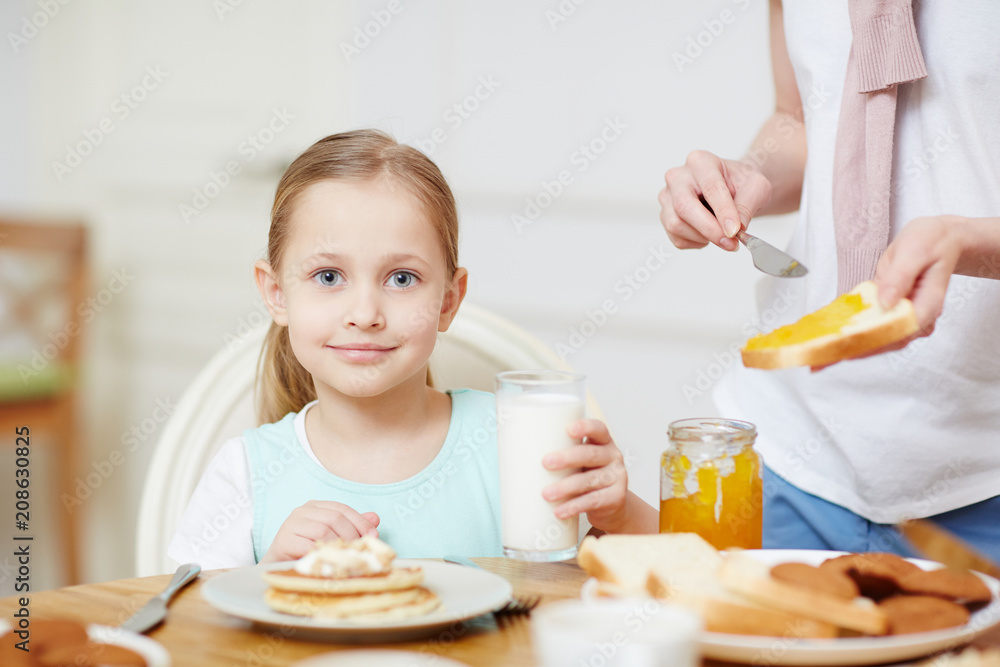 Pretty little girl with glass of milk looking at camera by served table