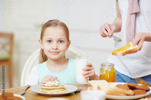 Pretty little girl with glass of milk looking at camera by served table