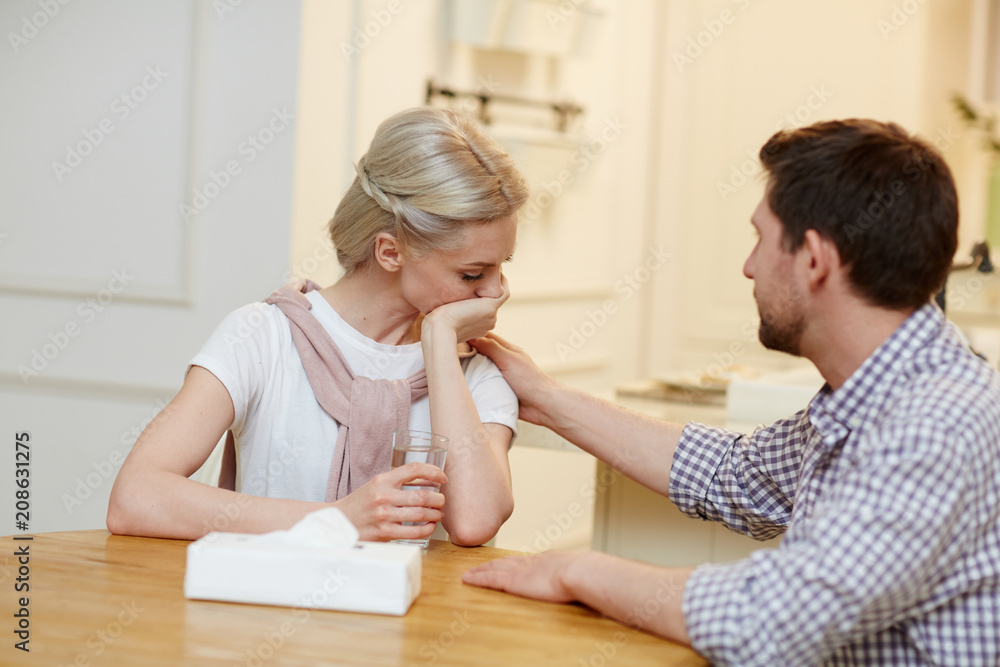 Unhappy woman with glass of water sitting by table while her husband comforting her Unhappy woman with glass of water sitting by table while her husband comforting her