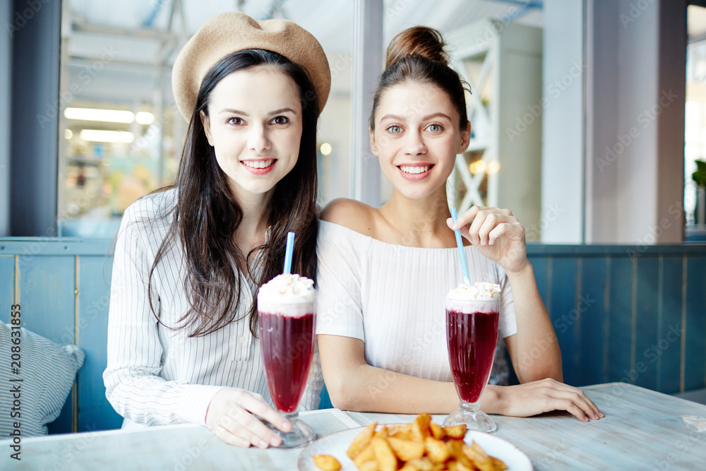 Happy young women in casualwear looking at you while having drinks in cafe Happy young women in casualwear looking at you while having drinks in cafe