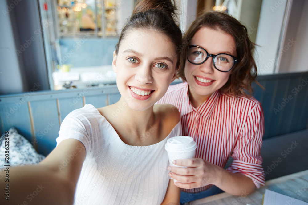 Two friendly girls with toothy smiles making selfie while relaxing in cafe Two friendly girls with toothy smiles making selfie while relaxing in cafe