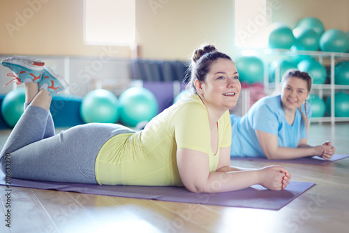 Two young chubby females lying on mats in fitness center and listening to instructor