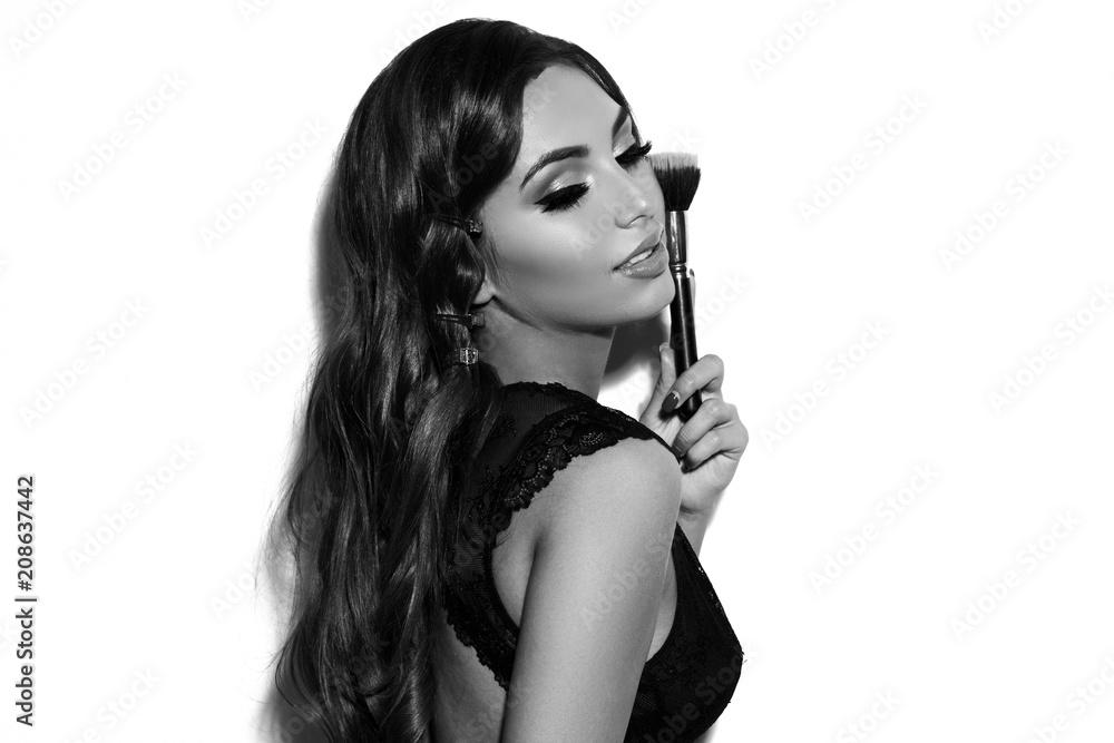 Obraz premium Young beautiful tanned woman in lave black bodysuit and blue jeans with long wavy brunette hair holding makeup brush and posing against white background