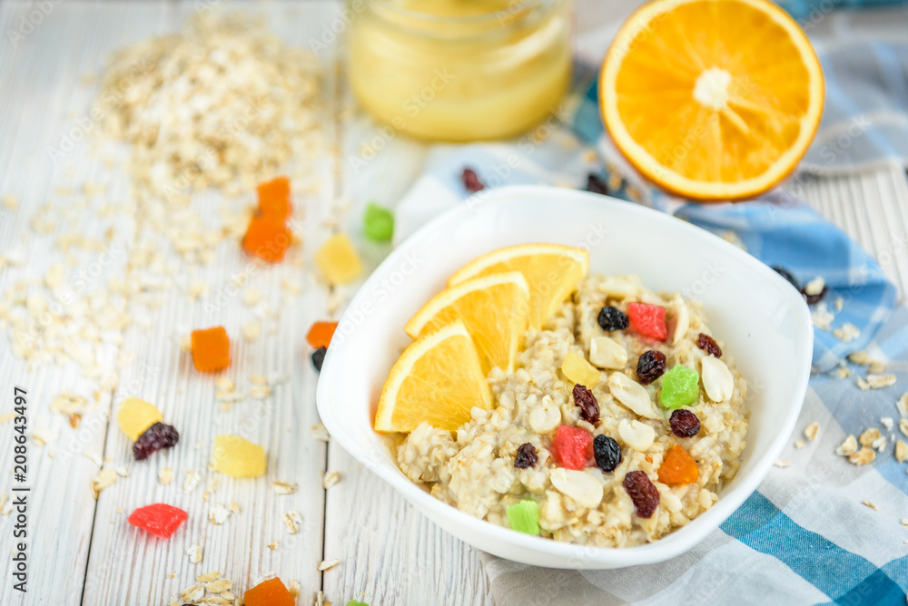 Homemade oatmeal porridge with honey, orange, raisin, peanut and candied fruit on white wooden background. Healthy breakfast.