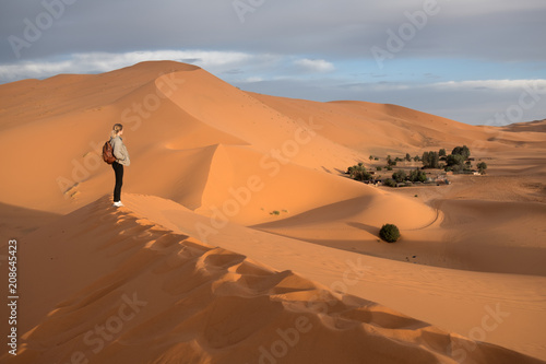 Girl with backpack standing at the top of a sand dune in Sahara desert, watching oasis in a distance during sunrise photo