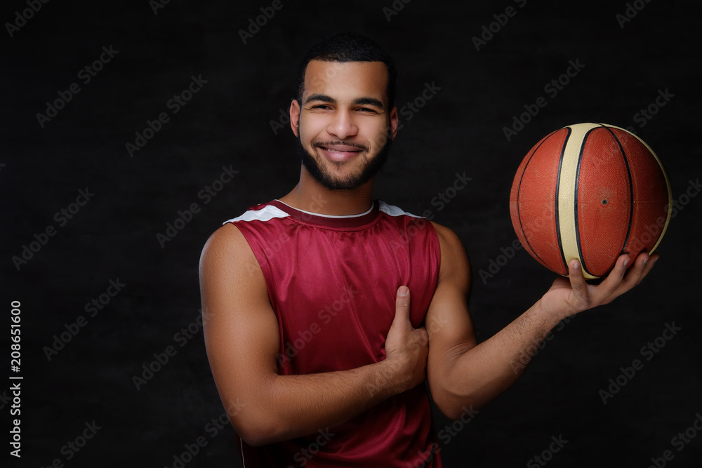 Smiling young African-American basketball player in sportswear isolated over dark background. 
