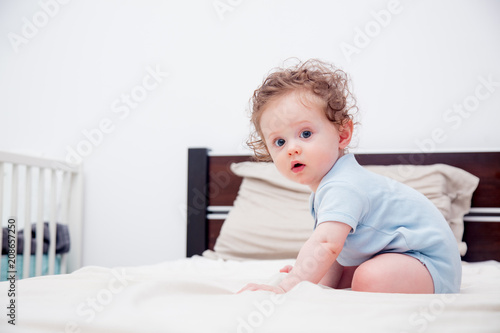 Curly little baby boy sitting on a bed in a bedroom.