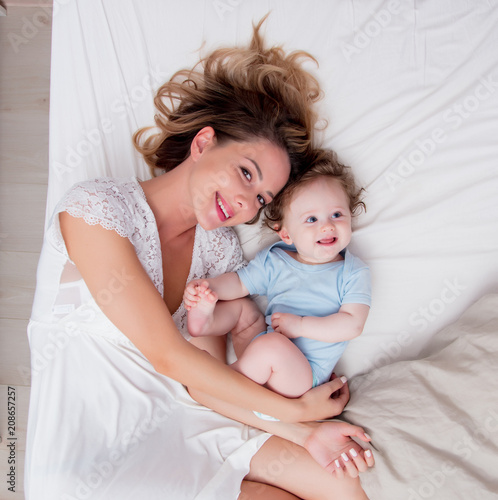 Young mother with her 7 month old little son dressed in pajamas are relaxing and playing in the bed at the weekend together, lazy morning, warm and cozy scene.