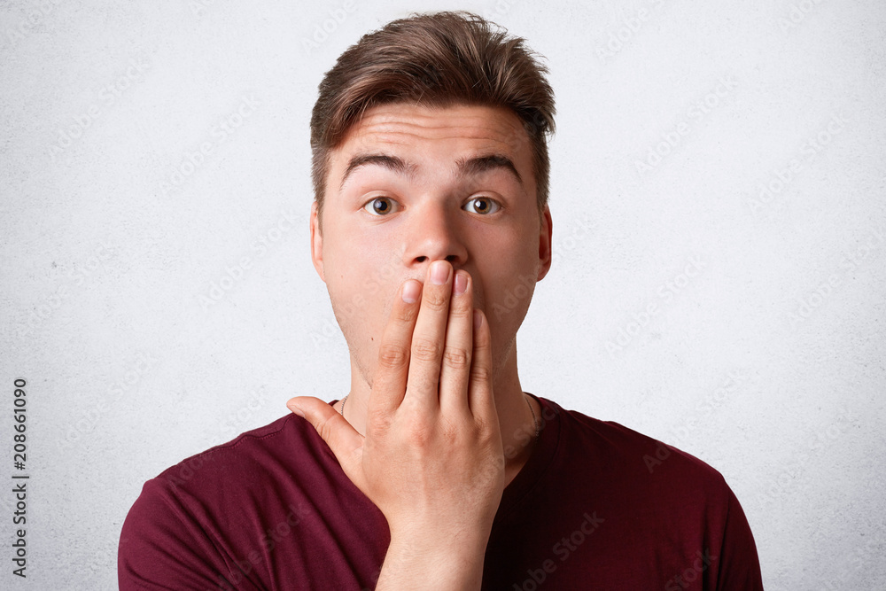 Headshot of stressful surprised male covers mouth and looks with astonishment, tries to be speechless, poses against white backround. Stunned handsome man expresses surprisement, recieves news Headshot of stressful surprised male covers mouth and looks with astonishment, tries to be speechless, poses against white backround. Stunned handsome man expresses surprisement, recieves news